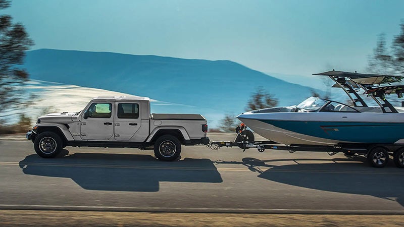 White Jeep Gladiator pickup truck towing a boat trailer along a scenic waterfront road.