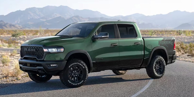 Dark green Ram Rebel pickup truck with off-road tires, parked on a road in a desert mountain landscape.