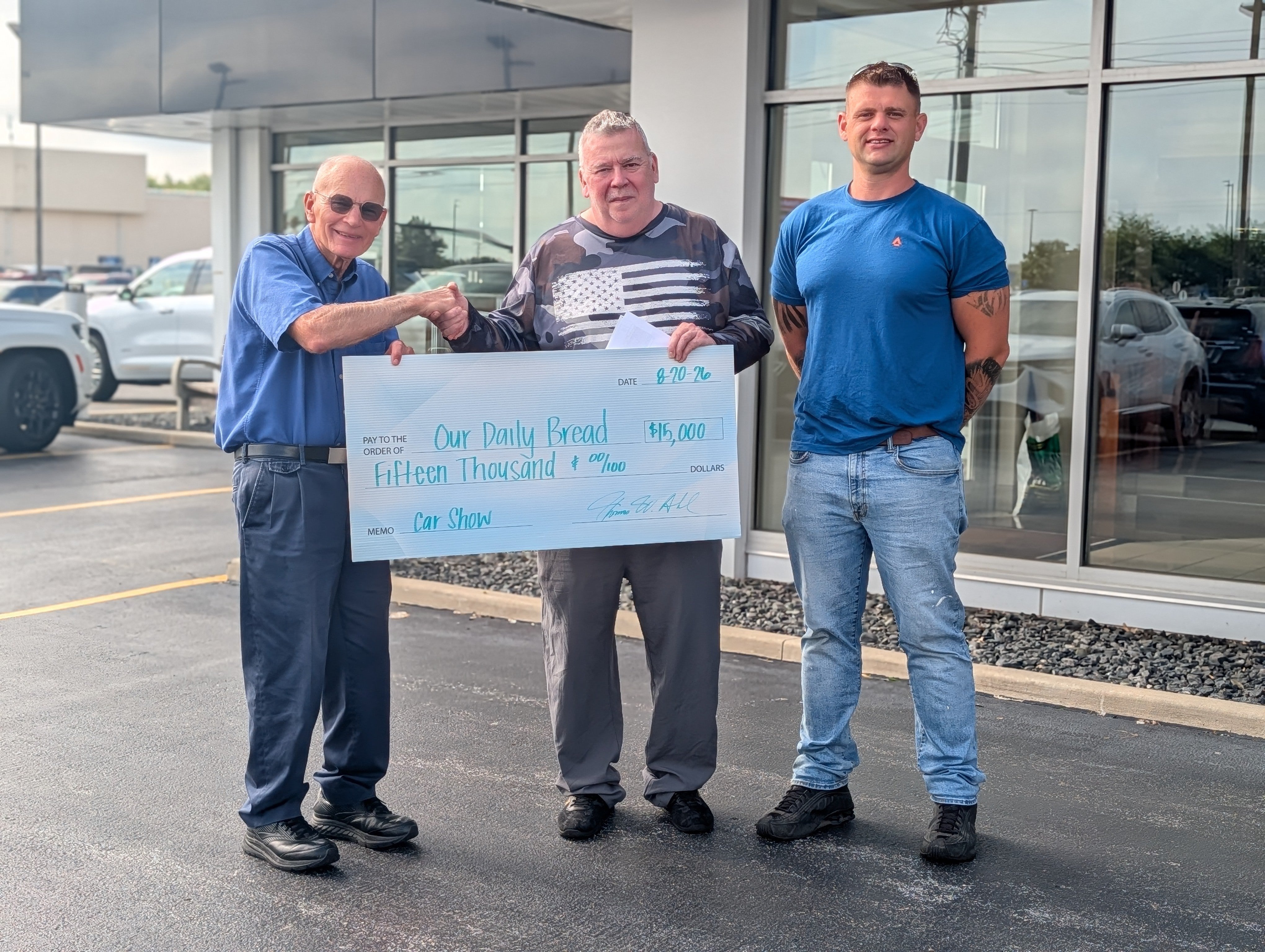 Three men pose outside holding an oversized $15,000 donation check made out to Our Daily Bread, with two shaking hands.