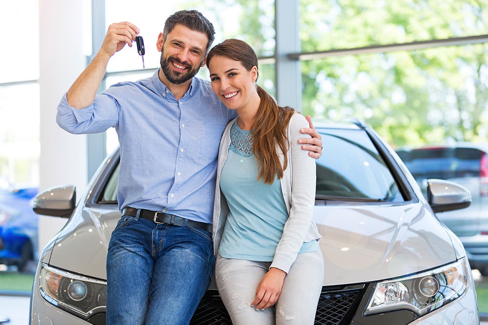 Smiling couple holding car keys, leaning on a new silver car in a dealership.