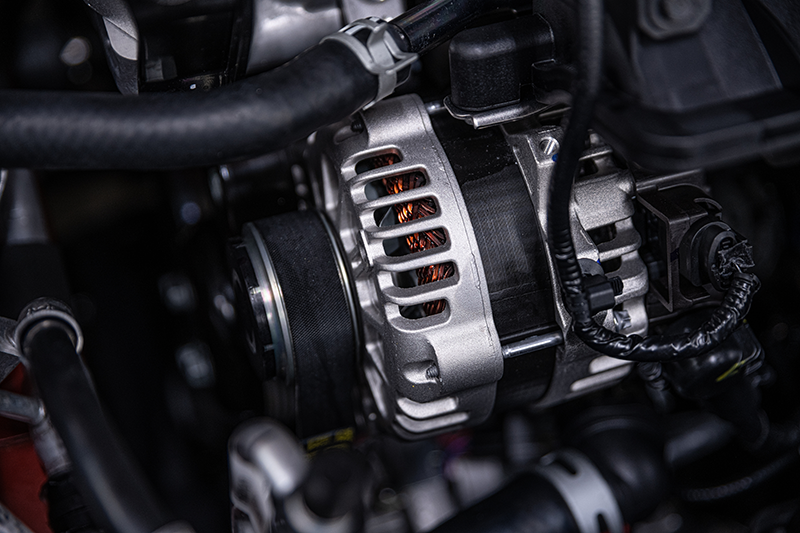 Close-up of a car engine's alternator, with a pulley and electrical wiring, surrounded by hoses.
