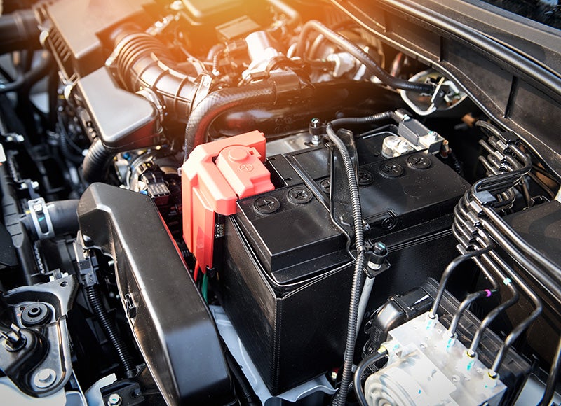 Automotive battery installed in a vehicle engine compartment, showing battery terminals, wiring