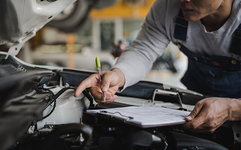 Mechanic inspecting an open car engine bay, holding a clipboard and pen.