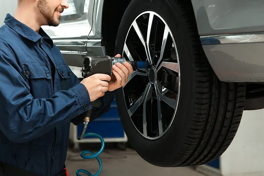 Mechanic in blue overalls using an impact wrench to work on a car's lifted wheel.